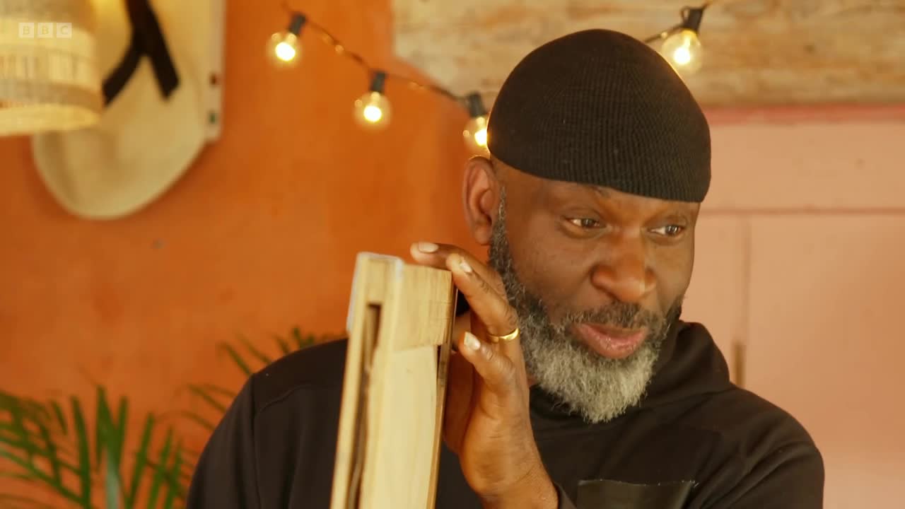 The man with a grey beard holds a wooden object to his ear, listening intently. String lights glow behind him against an orange wall, a familiar sight on BBC Two.