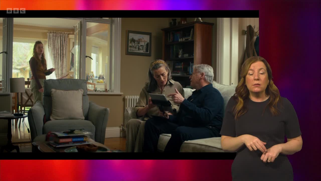 A woman stands by a window, gesturing as she speaks. On a sofa, an older couple looks at a tablet together.