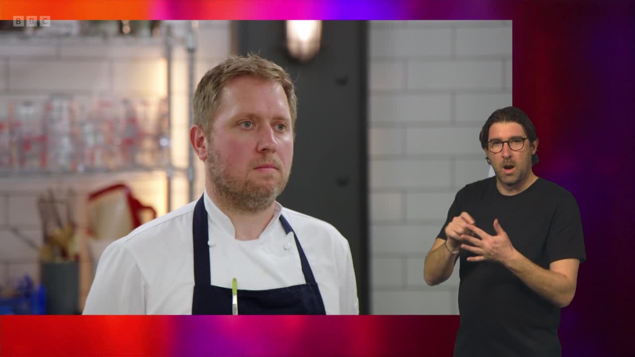 A chef stands in a kitchen, his expression serious as he looks off-camera. To his right, a man in a black t-shirt and glasses gestures emphatically, his mouth open in speech. The BBC Two logo is visible in the corner of the screen.