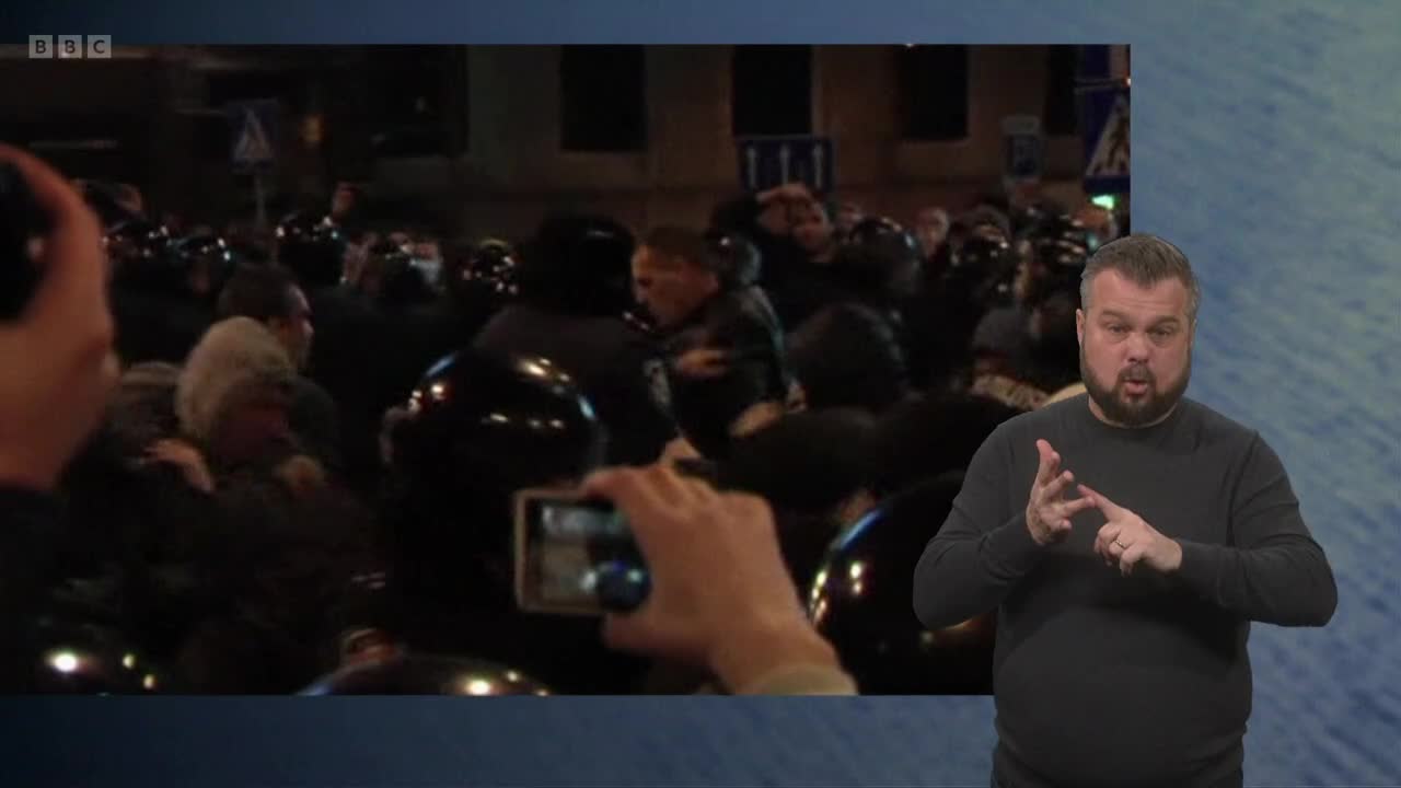 A crowd of people, some in black helmets, are gathered, and a phone is held up to record them. A man in a dark sweater signs along with the broadcast on BBC Two.