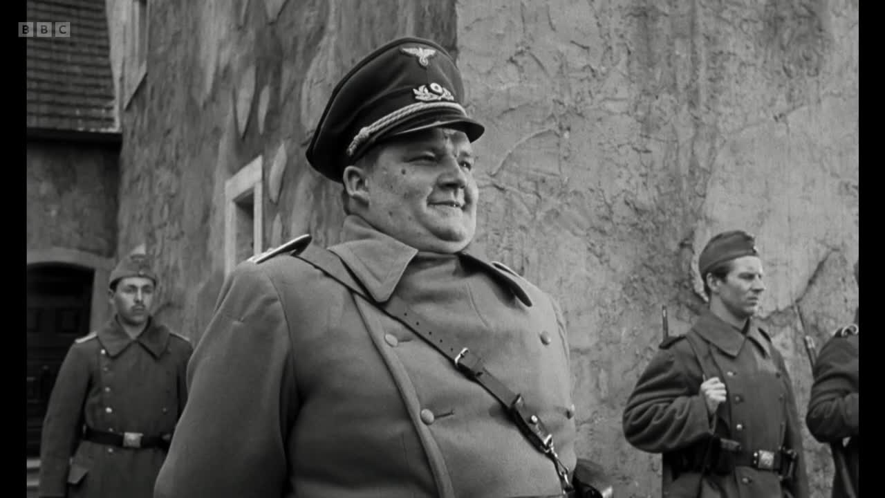 A portly officer in a peaked cap stands with a slight smile, his uniform immaculate. Behind him, two younger soldiers in similar attire stand at attention, their faces impassive.