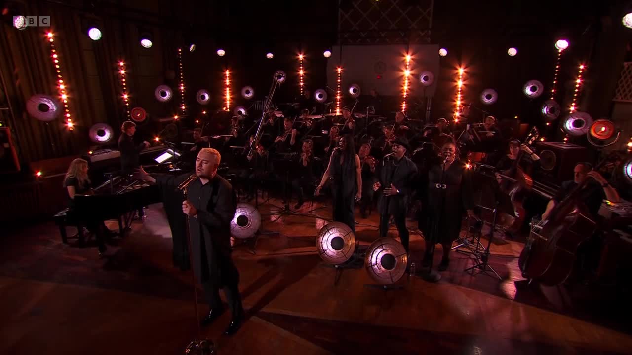 A singer in a black robe stands center stage, microphone in hand, as an orchestra plays behind him. The BBC Two logo is visible in the top left corner.