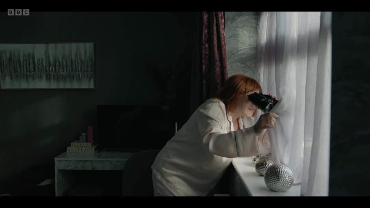 A woman with red hair peers through a gap in the curtains with binoculars. She's wearing pale pajamas and stands by a windowsill.