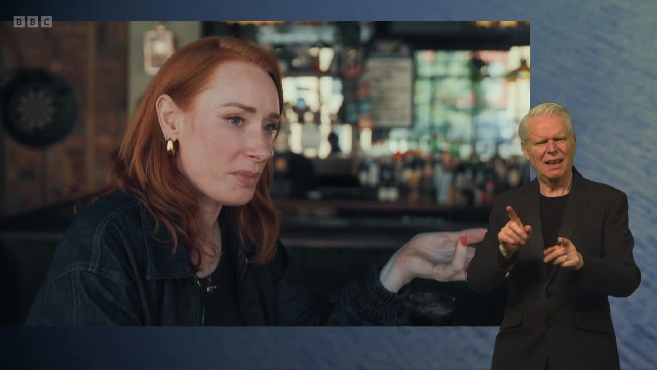 A woman with red hair looks intently to her left, her hand gesturing slightly. Beside her, a man in a suit signs with his hands, his expression animated. The BBC Two logo is visible in the corner.