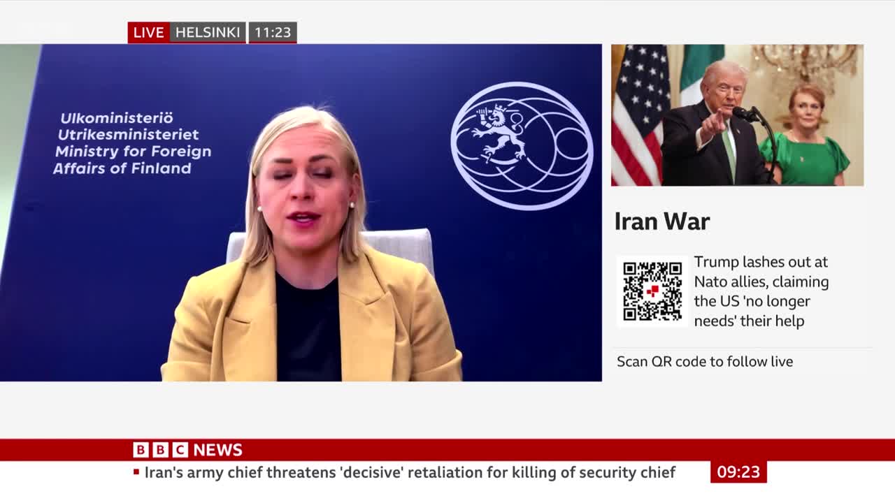 A woman in a tan jacket speaks directly to the camera, her Finnish Ministry for Foreign Affairs backdrop behind her. To the right, a smaller screen shows Donald Trump pointing, with text about "Iran War" and "Trump lashes out at Nato allies."