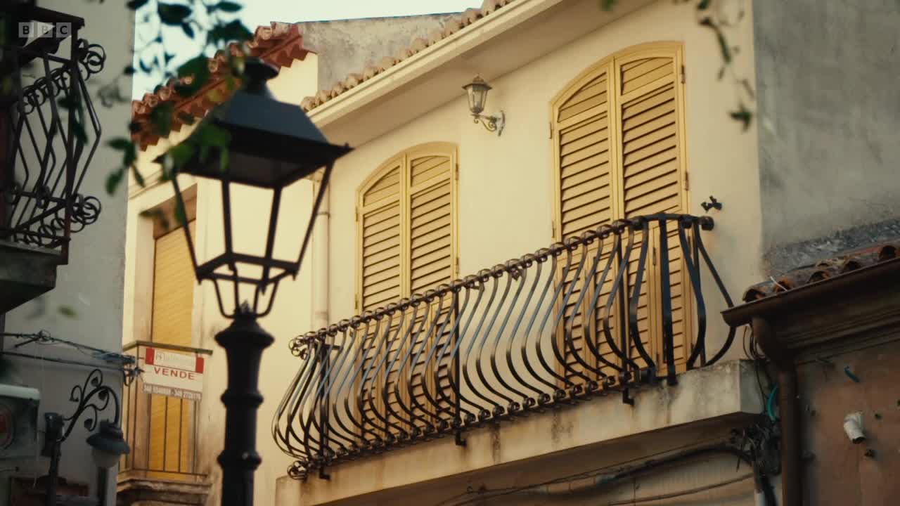 A red "VENDE" sign hangs on the side of a building, its shutters closed. A wrought-iron balcony curves gracefully in front of the windows, overlooking a narrow street.
