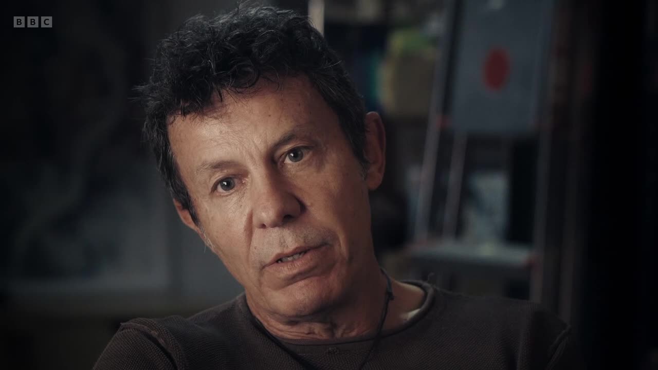 A man with dark, curly hair looks directly at me, his face angled slightly. Behind him, out of focus, are shelves holding various objects, including what looks like a framed picture with a red circle.