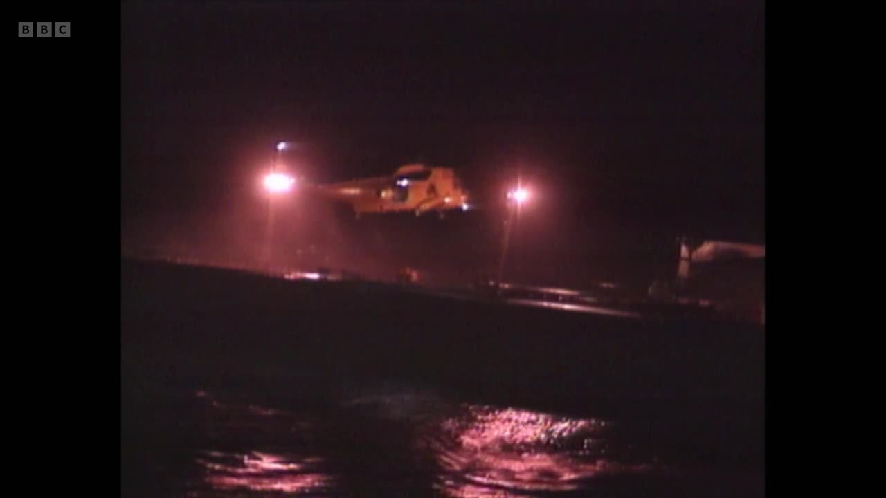 A yellow helicopter hovers low over a dark, choppy sea, its searchlights cutting through the night. The BBC Two logo is visible in the corner, suggesting this is part of a broadcast from the United Kingdom.