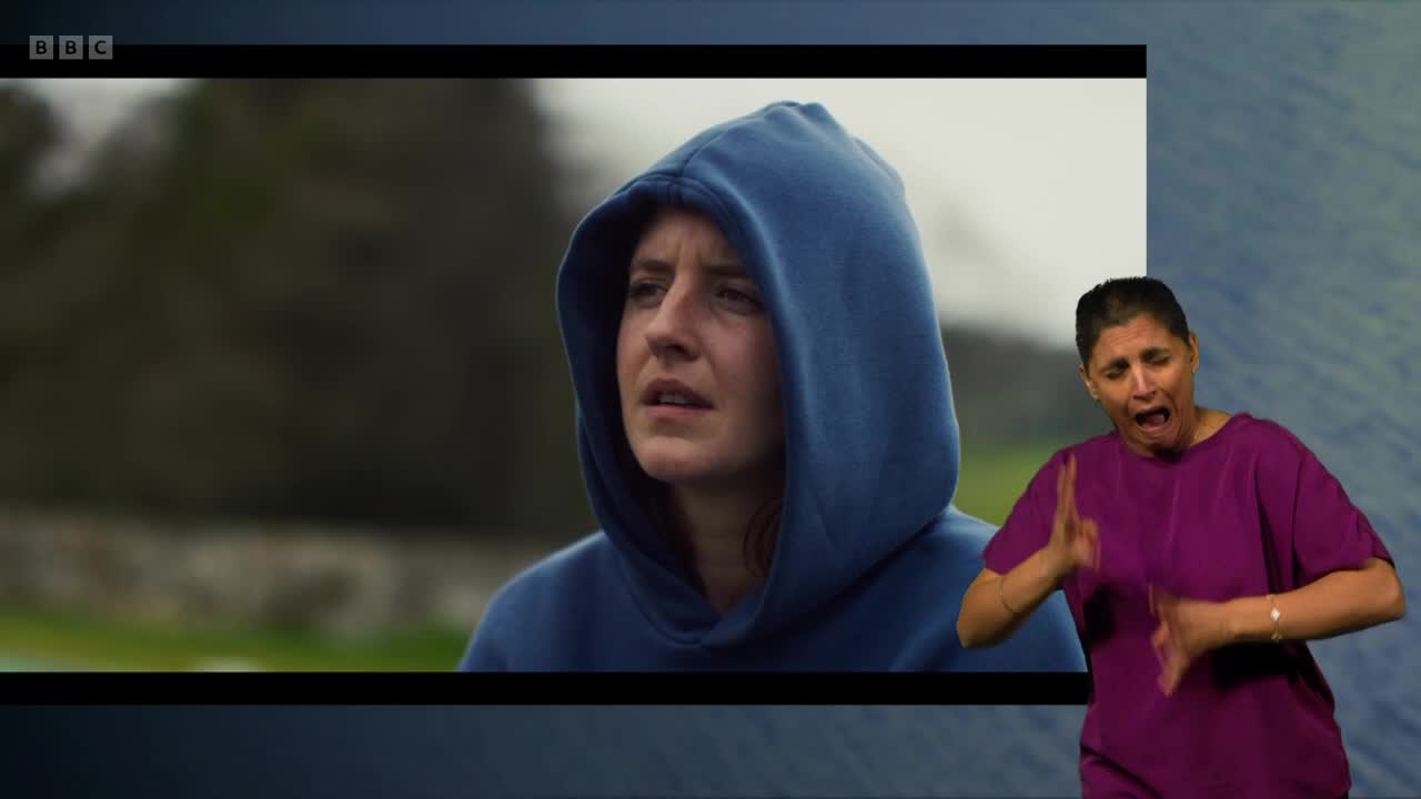 A young woman in a blue hoodie looks off-screen with a concerned expression. Beside her, a woman in a purple top signs with animated gestures. The BBC Two logo is visible in the corner.