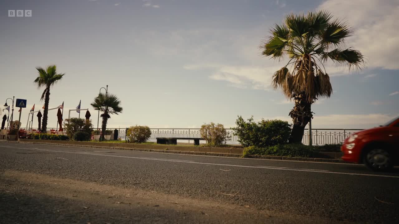 A red car drives past palm trees lining a coastal road. The BBC Two logo is visible in the top left corner.