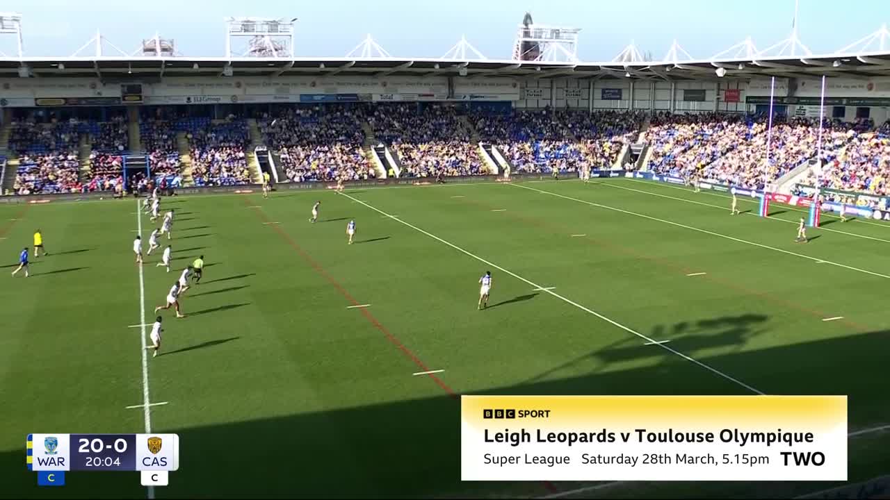 Players in white jerseys are spread across the pitch, some running with the ball. The crowd in the stands is a sea of blue and yellow, watching the Super League match.