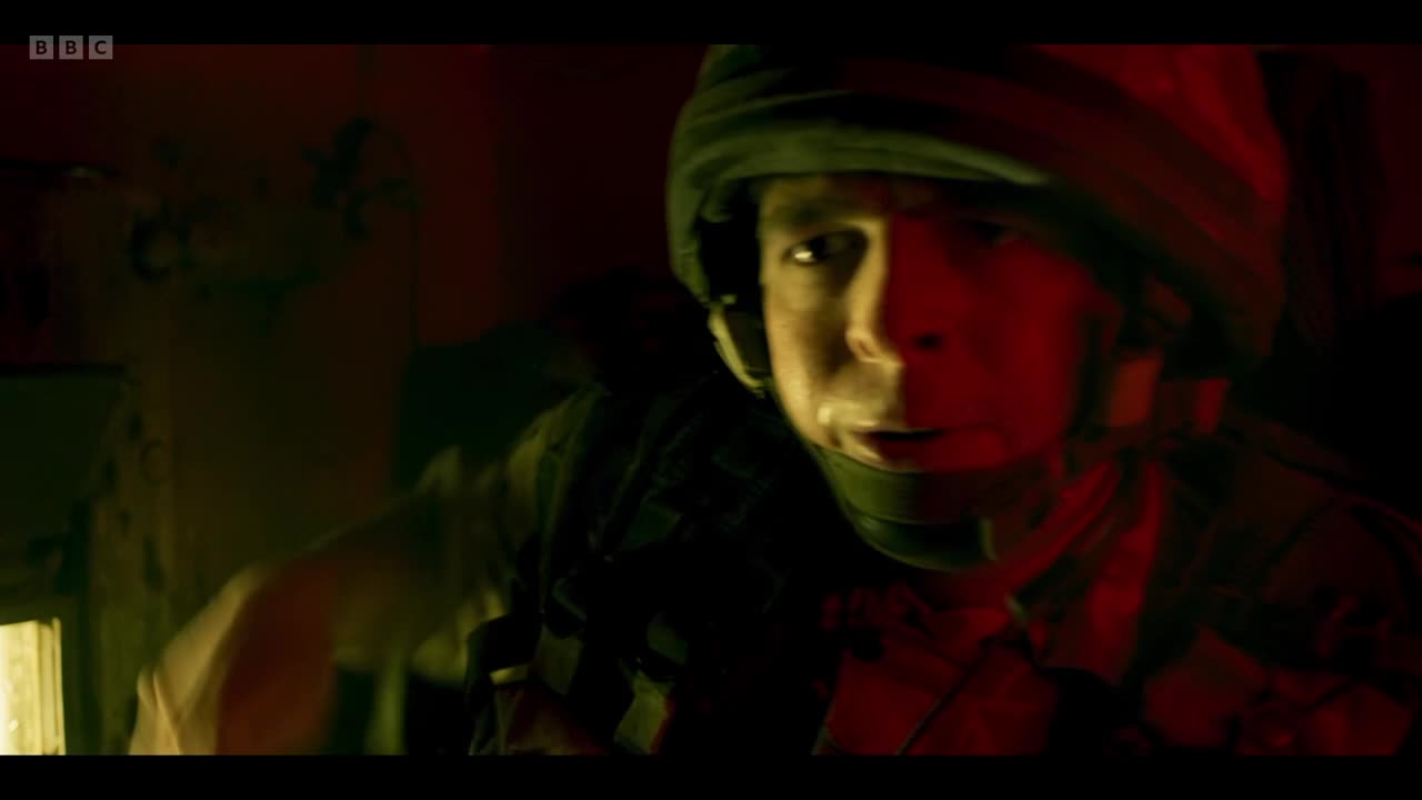 A young soldier, helmeted and clad in camouflage, strains forward in the dim, red-tinged interior of a vehicle, his face etched with a mixture of fear and determination. The BBC Two logo is visible in the corner, hinting at a dramatic scene unfolding in the United Kingdom.