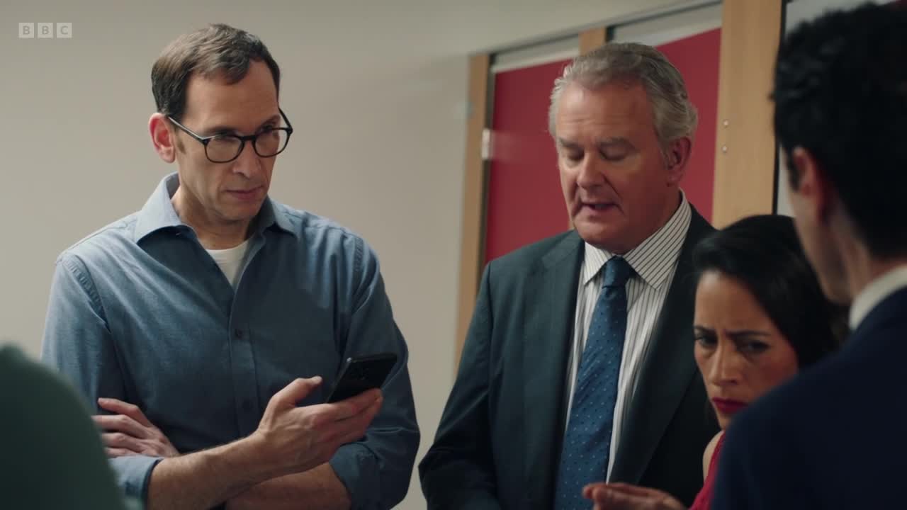 A man in glasses holds up a phone, his brow furrowed. Beside him, a man in a suit speaks intently, his gaze directed downwards.