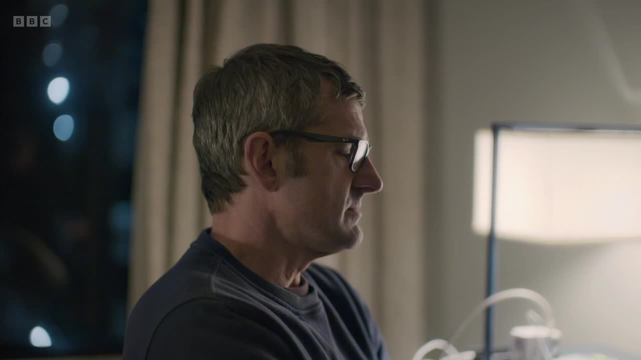 A man in glasses sits looking out a window at the blurred lights of a city at night. He wears a dark blue jumper, and a lamp is visible behind him.