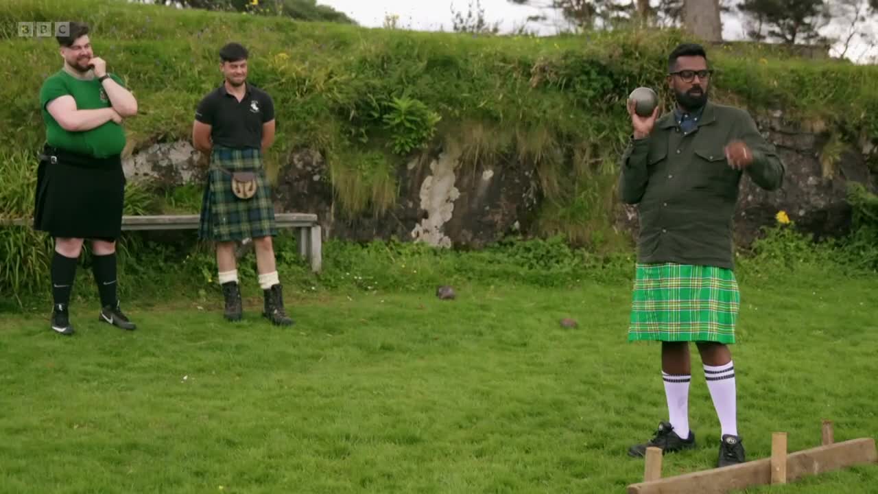 A man in a green kilt is about to throw a heavy stone. Two other men, also in kilts, watch him from the side. This is part of a segment for BBC Two.