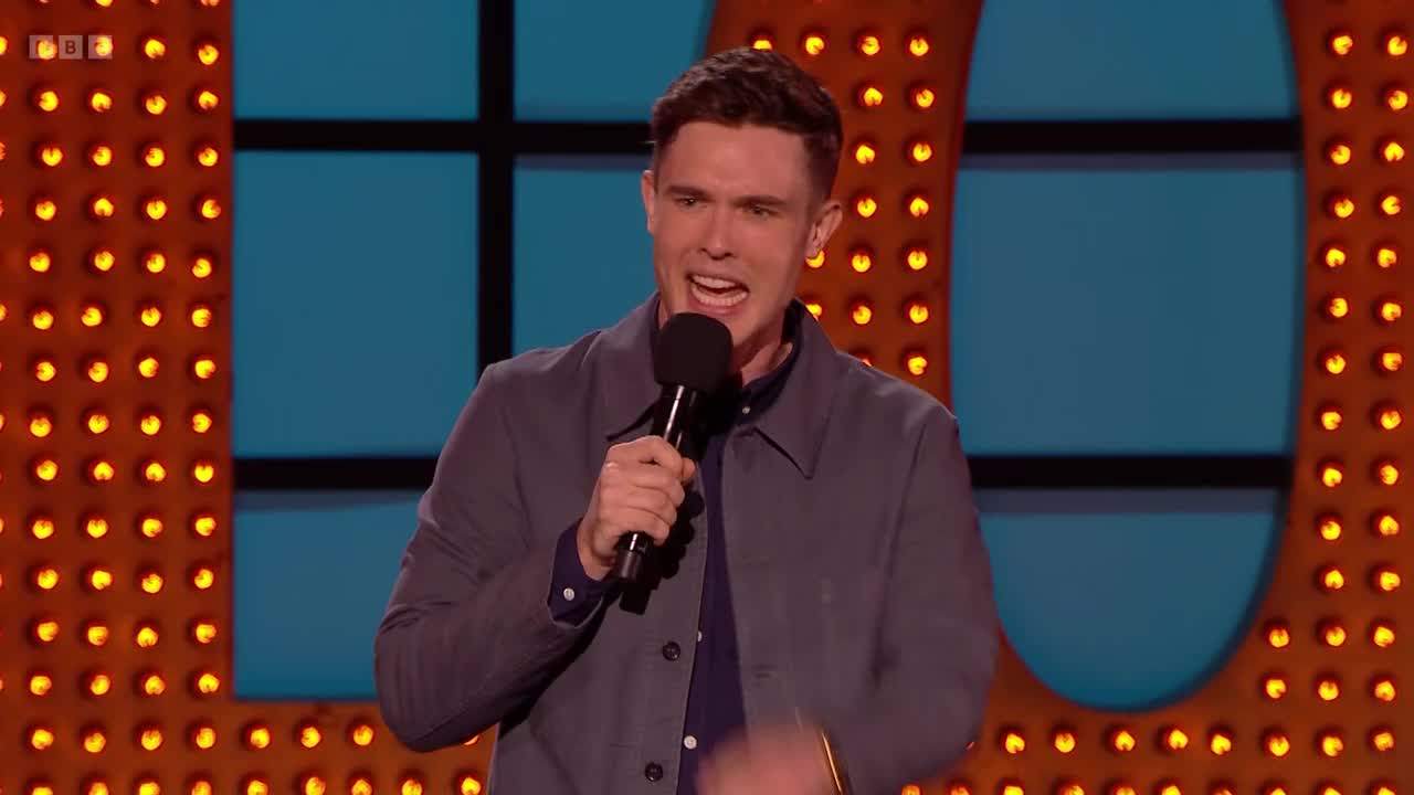 A comedian grips the microphone, his mouth open mid-sentence on the BBC Two stage. He's framed by rows of warm, orange lights.