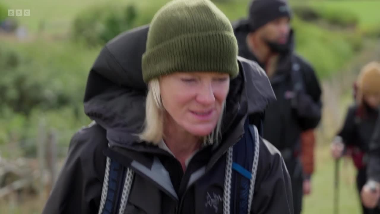 A woman in a green beanie and a dark jacket walks forward, her backpack heavy on her shoulders. Behind her, two other figures, also with backpacks, follow along a grassy path.