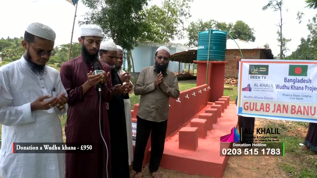 A group of men, some wearing white caps, stand in a field near a newly-built washing area. One man speaks into a microphone, while others have their hands raised in prayer.
