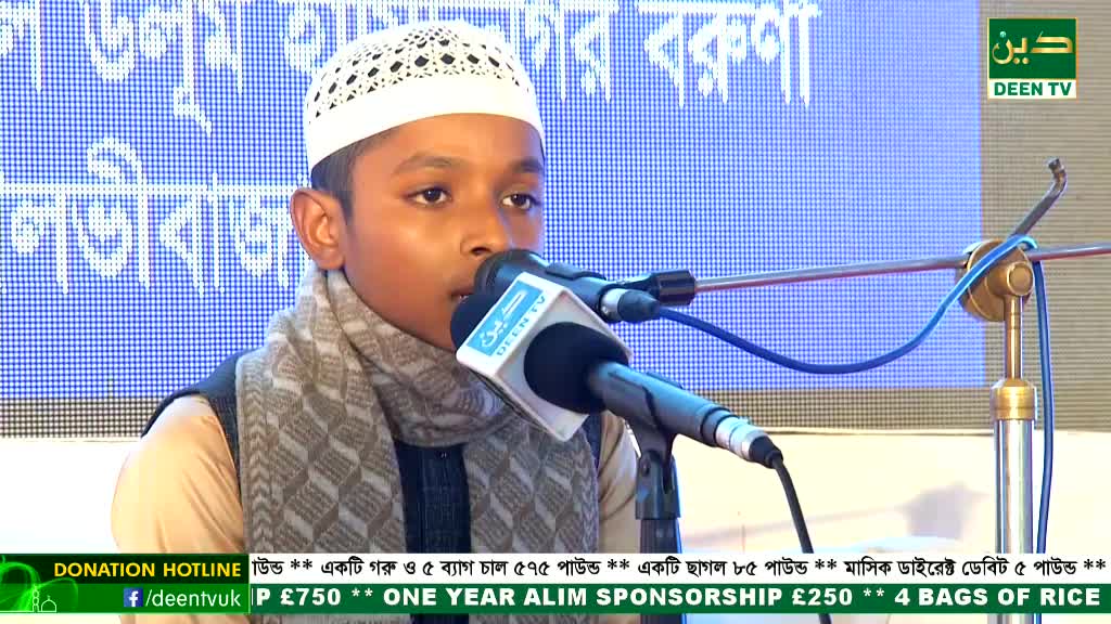 A young boy, wearing a white cap and scarf, speaks into a microphone, his voice amplified by Deen TV. Behind him, a blue backdrop displays Bengali script and contact information for donations.
