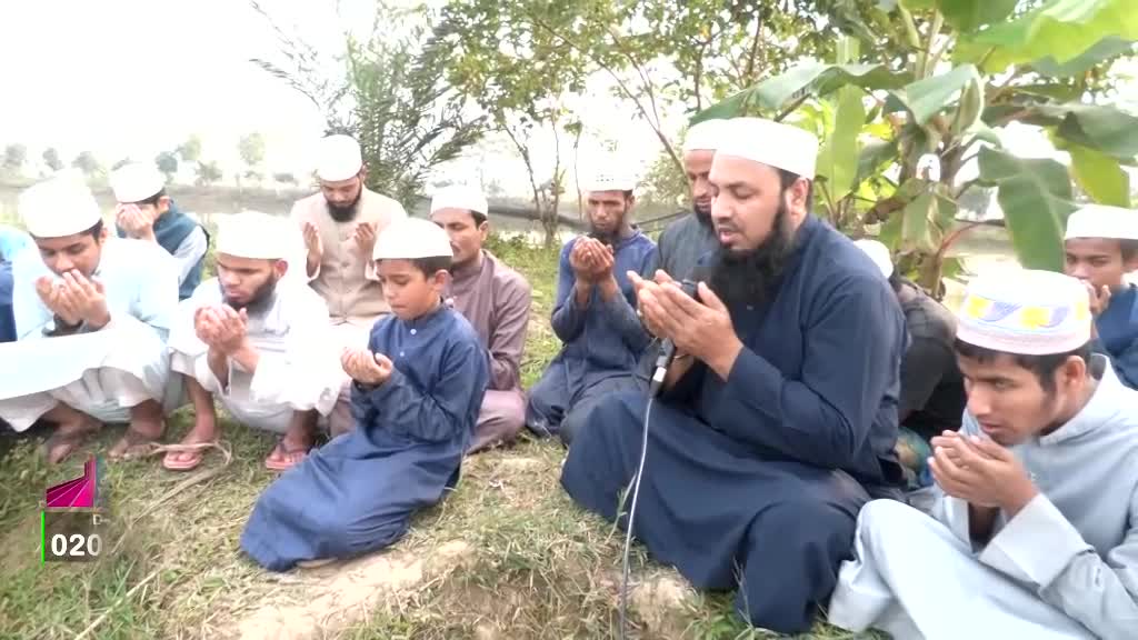 Men and boys, all wearing white caps, are kneeling in a circle outdoors, their hands raised in prayer. A man with a long beard and a microphone is leading the prayer, likely for a Deen TV broadcast.
