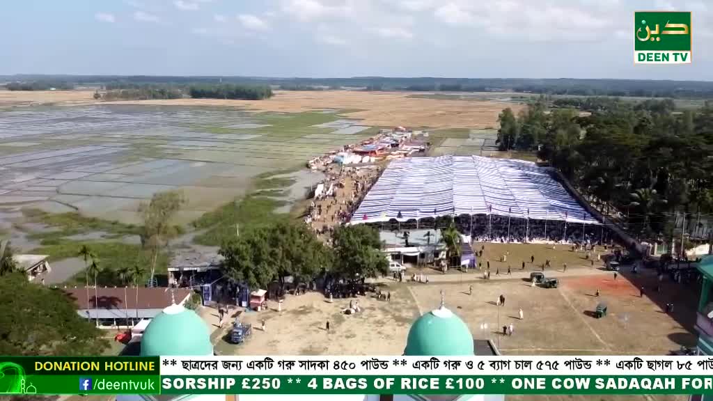 A large crowd gathers under a massive white tent, while Deen TV's donation hotline scrolls across the screen. Beyond the event, flooded fields stretch towards a distant tree line.
