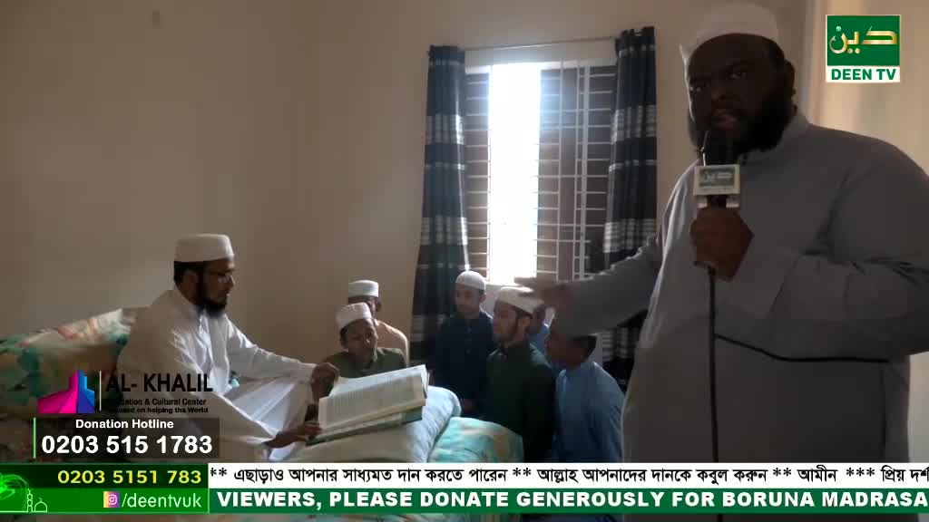A man in a light grey robe speaks into a microphone, gesturing towards a group of young students. They're gathered around a man reading from a large book, likely in a classroom setting.
