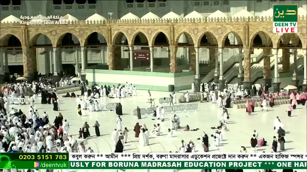 People in white garments move across the vast, pale stone courtyard, some kneeling and others walking towards the arched colonnades. The live broadcast from Deen TV in the United Kingdom shows the scene with a banner at the bottom.

