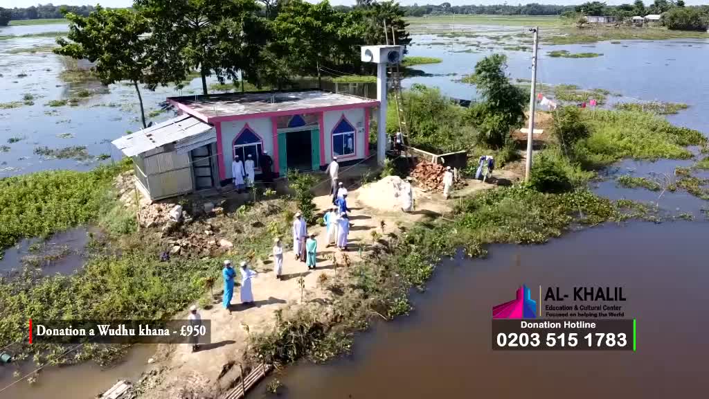 People in white robes and caps walk toward a small building with a pink and white facade, possibly a mosque. The structure sits on a small island surrounded by water, with a few others working nearby.
