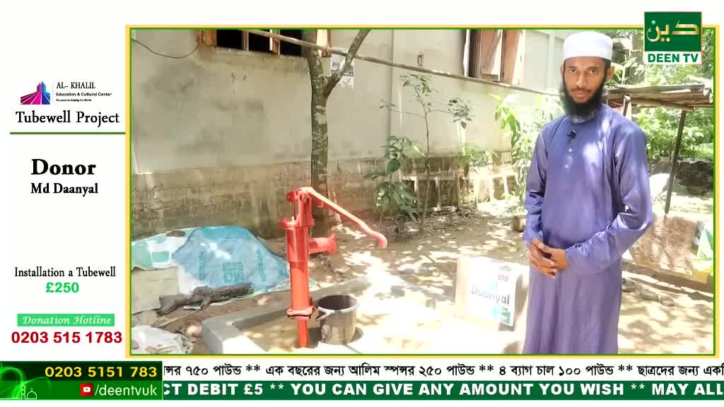 A man in a purple tunic stands near a red water pump, likely for a Deen TV project.  Behind him, a concrete structure and a small tree are visible, as the pump is set up outdoors.
