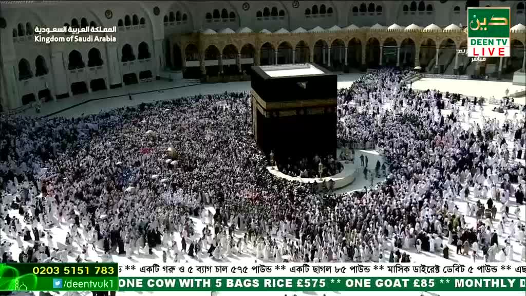 A massive crowd in white garments surrounds a black cube, all moving in a circular pattern. The scene is broadcast live on Deen TV, with details of charitable donations scrolling at the bottom.
