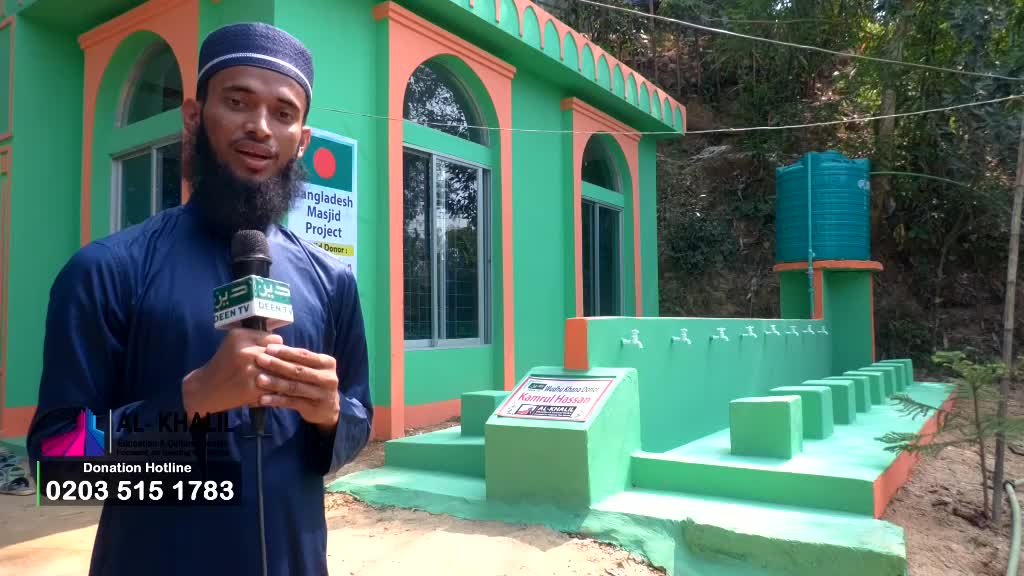 A man with a beard, holding a microphone from Deen TV, stands in front of a green and orange building. Behind him, a water tank sits atop a structure with multiple taps.
