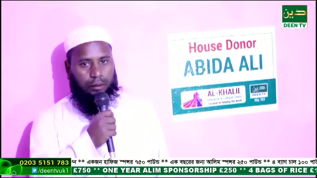 A man in a white robe and cap speaks into a microphone, facing the camera. Behind him, a sign on the wall for Deen TV in the United Kingdom promotes a house donation and other sponsorships.

