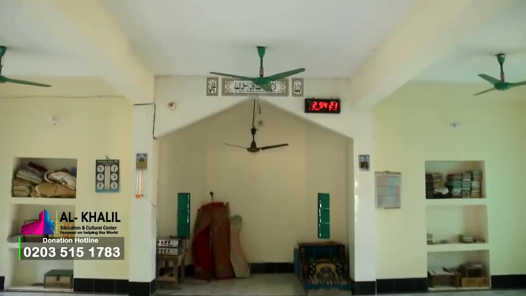 Three green ceiling fans slowly spin, stirring the air in a simple, light-colored room. Above the arched doorway, a digital clock displays the time, and a banner for Al-Khalil Education & Cultural Center is visible.
