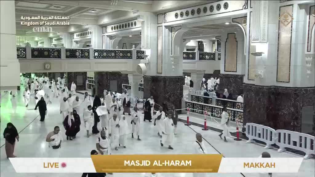 People in white garments are moving through a vast, ornate hall. The scene is being broadcast live from Masjid Al-Haram in Makkah, as seen on Eman Channel.
