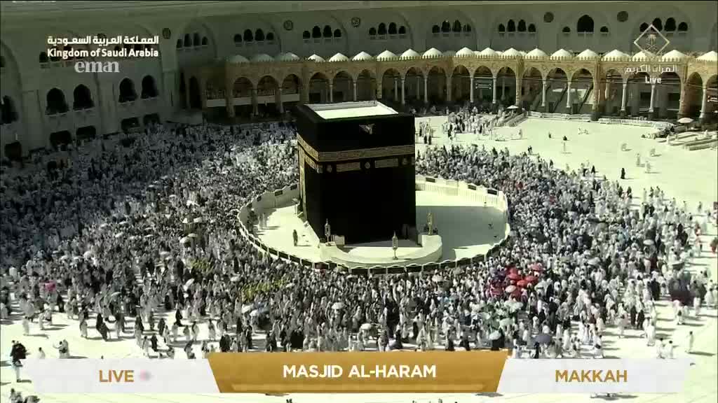 A sea of people in white garments flows around the black cube, some carrying umbrellas. The scene is being broadcast live from Masjid Al-Haram, as seen on Eman Channel.
