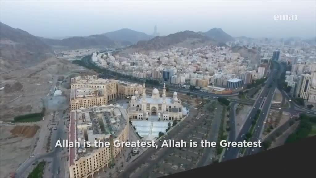 A large, ornate mosque sits at the center of a sprawling city, its white facade contrasting with the surrounding beige buildings. The Eman Channel logo is visible in the top right corner, and the words "Allah is the Greatest" scroll across the screen.

