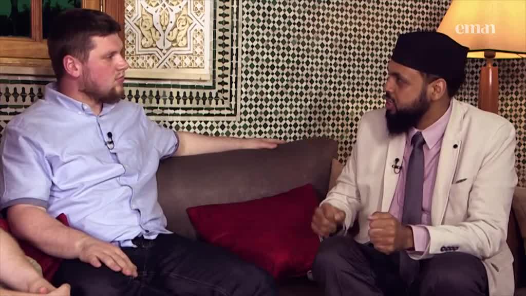 On a set for Eman Channel, two men are seated on a couch, engaged in conversation. The man in the black cap gestures emphatically while speaking.
