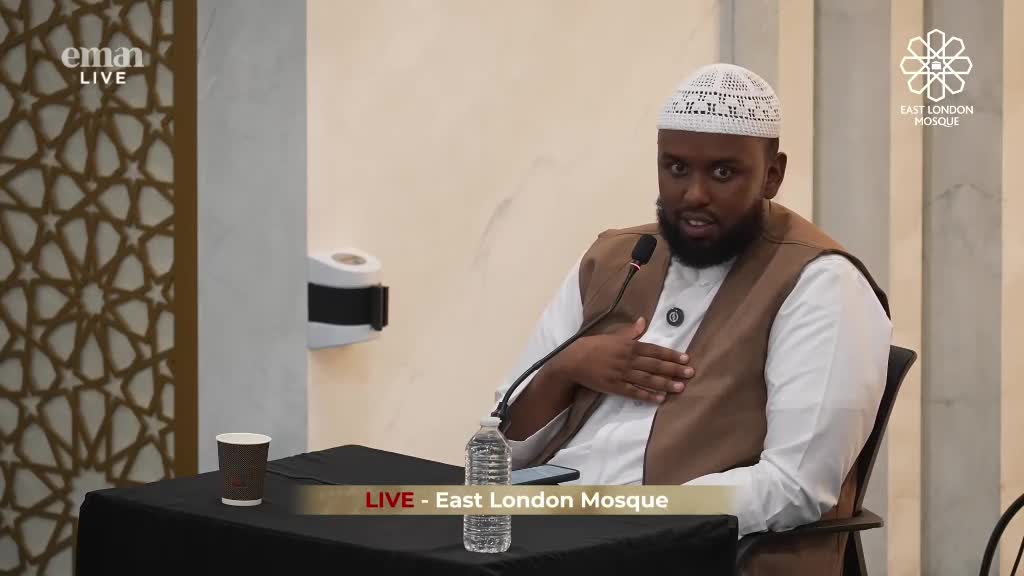 A man in a white cap and brown vest speaks into a microphone, gesturing with his right hand. The live broadcast from the East London Mosque is clearly visible on the Eman Channel.
