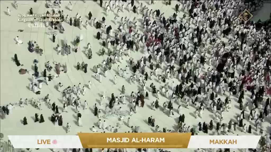 A sea of people, mostly in white, flows across the vast, light-colored space. The Eman Channel broadcast from the United Kingdom is showing the live feed from Masjid al-Haram in Makkah.
