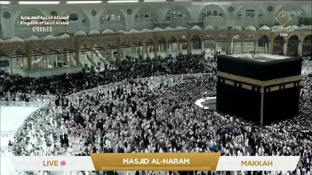 A dense crowd of people, mostly dressed in white, circles the black cube structure. The scene is being broadcast live from Masjid al-Haram, with the Eman Channel logo visible.

