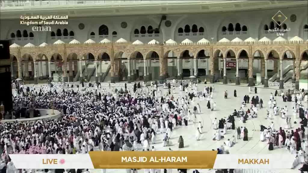 A vast crowd in white garments moves slowly around a central structure, the scene broadcast live from Masjid al-Haram on Eman Channel. The architecture is grand, with arched walkways and ornate details, and the atmosphere feels solemn and reverent.
