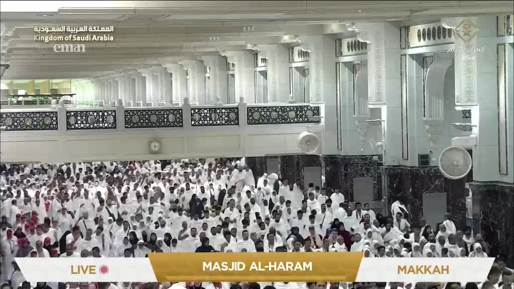 A dense crowd of people in white garments fills the vast hall, moving slowly. Overhead, the Eman Channel logo and the words "Masjid Al-Haram" are visible.
