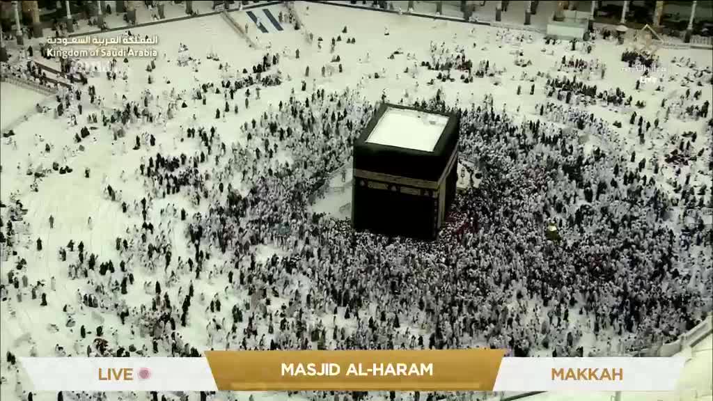 A dense crowd of people, mainly in white, flows around a black, cube-shaped structure. The scene is being broadcast live from Masjid al-Haram in Makkah, as seen on the Eman Channel.
