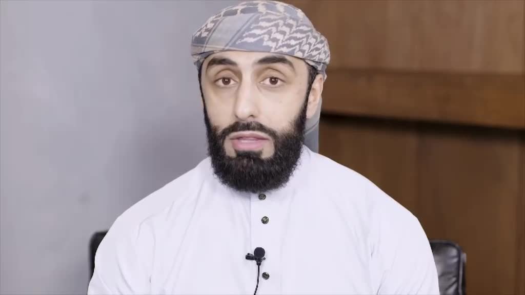 A man with a full beard and a patterned head covering speaks directly into a small microphone. He's wearing a white tunic, and the background is a simple wooden panel.
