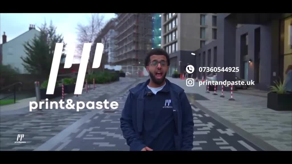 The man in the blue jacket stands on a paved walkway, speaking directly to the camera. Behind him, modern buildings and construction scaffolding frame the shot, likely somewhere in the United Kingdom.
