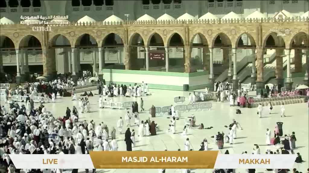 Pilgrims in white robes are moving across the vast courtyard, some praying and others walking purposefully. The scene is broadcast live from Masjid al-Haram in Makkah, as seen on Eman Channel.
