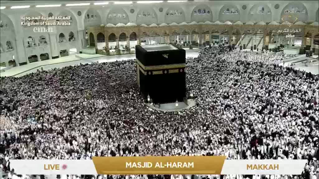 A vast crowd surrounds the Kaaba, a black cube-shaped structure. People are moving in a circular pattern, a sea of white garments and faces.

