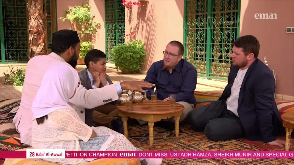 A group sits on cushions, gathered around a small table with glasses and tea pots. The man in the white robe gestures, his hand hovering over the table as he speaks.

