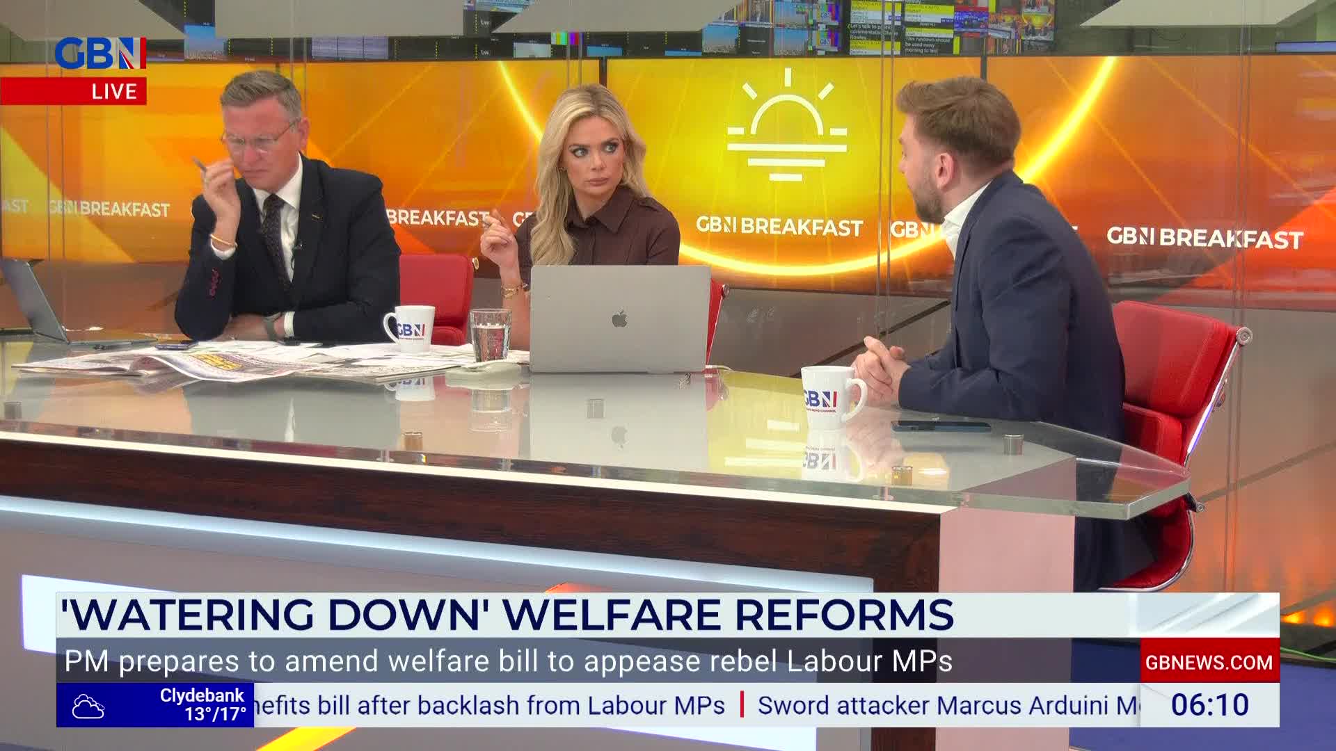 Three people are seated at a desk, discussing something on GB News' "Breakfast" show. The man on the left is holding a pen to his face, while the woman in the middle gestures with her finger.
