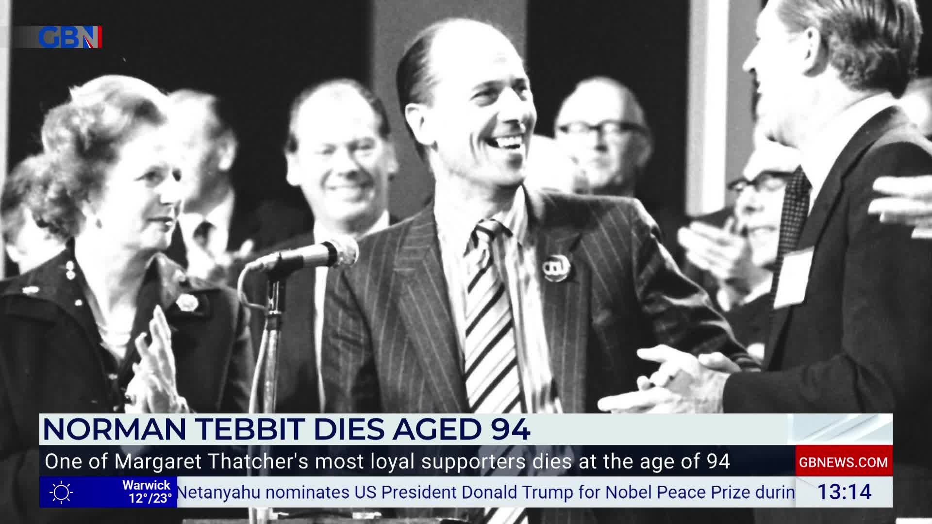 Norman Tebbit, smiling broadly, shakes hands with a man in a dark suit. Margaret Thatcher stands nearby, clapping, as the scene unfolds on the GB News broadcast.
